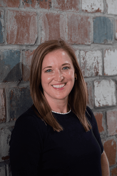 Headshot photo of woman with brown hair and blue sweater standing in front of a brick wall